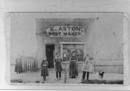 Four people stand outside of a shop with a sign E Aston Boot Maker