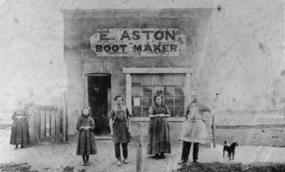 Four people stand outside of a shop with a sign E Aston Boot Maker