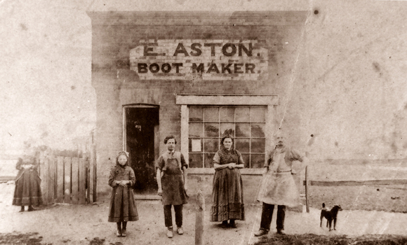 Four people stand outside of a shop with a sign E Aston Boot Maker