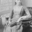 Tilly Aston as a young girl sitting in a chair with a handbag in her lap