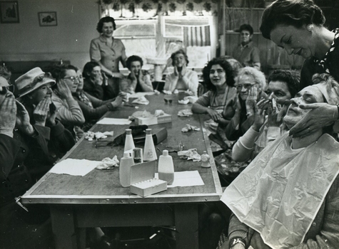 Long table of ladies rub lotion into their faces whilst one in the foreground receives a facial massage