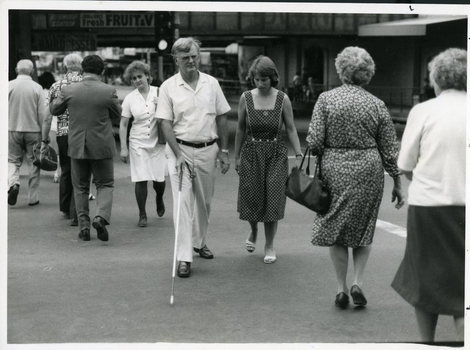 Man makes road crossing with cane whilst woman watches