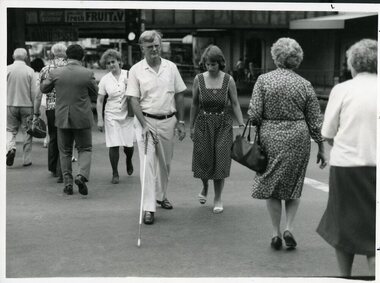 Man makes road crossing with cane whilst woman watches