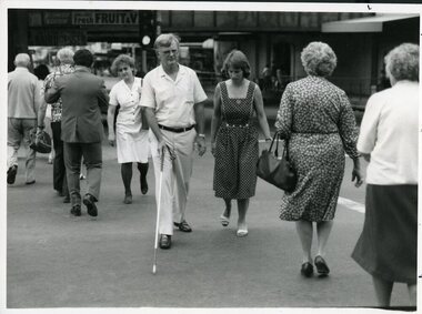 Man makes road crossing with cane whilst woman watches