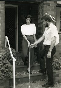 Man lightly lifts cane as woman descends steps from house.