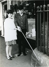 Woman raising white cane man is holding to make it touch a guttering