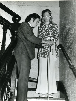 Blindfolded woman holds white cane as man tries to assist her down the stairs