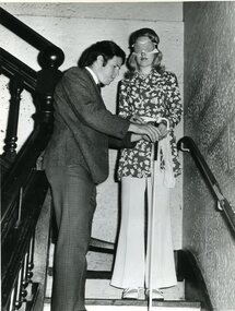 Blindfolded woman holds white cane as man tries to assist her down the stairs