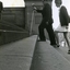 Man holds long white cane above steps as he ascends, watched by instructor