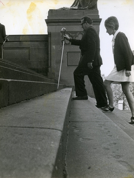 Man holds long white cane above steps as he ascends, watched by instructor