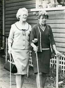 Two women using their white canes and a handrail as they walk along a path