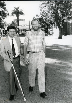Two men walk across a park, one with a cane and the other partially guiding