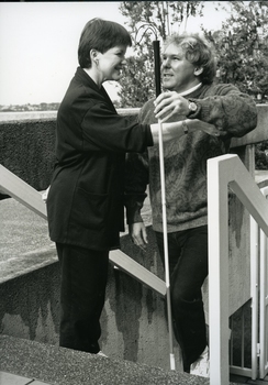 Man ascending stairs with white cane with woman at top with her hand under his elbow