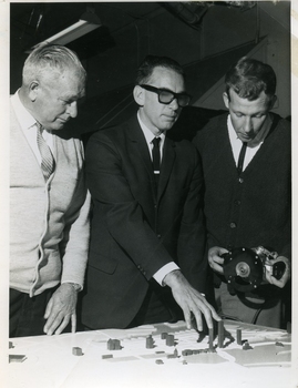 Man in suit touches the tops of buildings on a 3D map with two others looking on