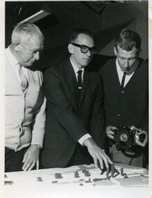 Man in suit touches the tops of buildings on a 3D map with two others looking on