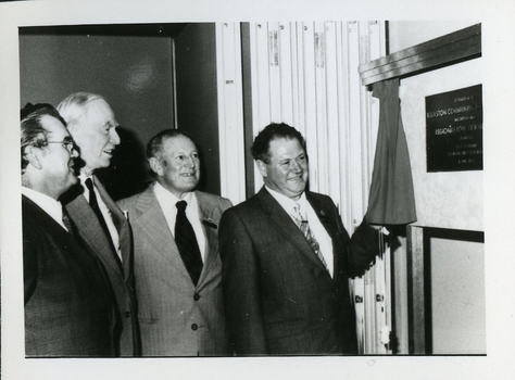 Three men stand and look at wall plaque with a fourth pulling aside the curtain that previously covered it