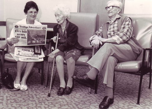 Lady reads Bendigo Advertiser to two elderly people seated in lounge at Mirridong