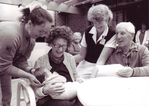 Elderly woman holds baby with  woman supporting baby's neck, nearby man laughing and nurse looking on