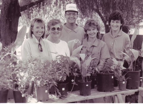 Five people stand smiling behind pots of plants
