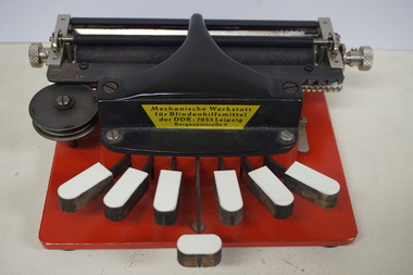 Metal braille machine with red base and 7 white wood keys