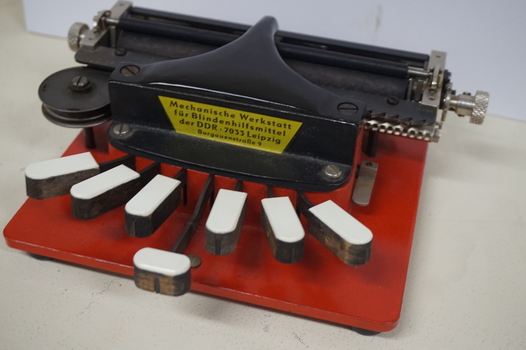 Metal braille machine with red base and 7 white wood keys