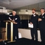 Three men stand on raised platform with one speaking at podium