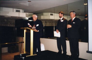 Three men stand on raised platform with one speaking at podium