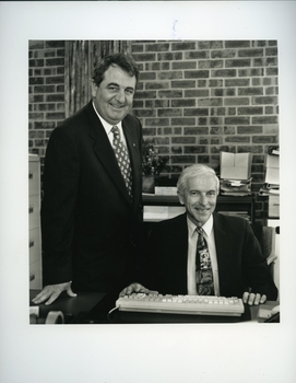 John Puttick stands beside seated Peter Lynam in an office