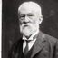 White haired older man wearing wire glasses looks to camera