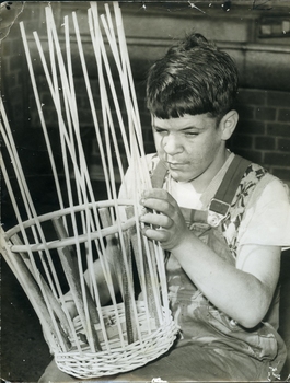 Teenage boy in overalls sits outside as he weaves a cane basket