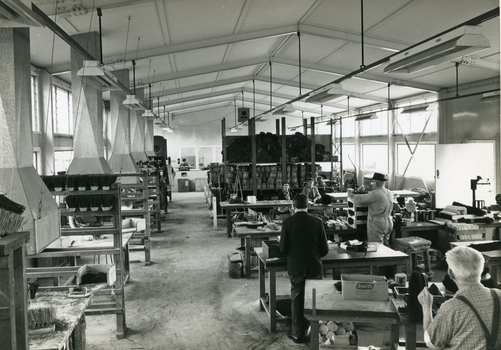 Men stand at workstations working on various types of brushes