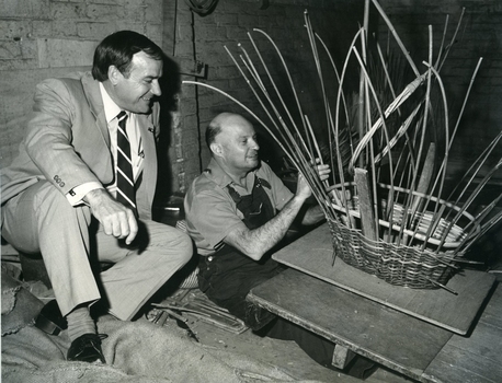 Man making a basket in the workshop whilst another man sits on sacks and watches