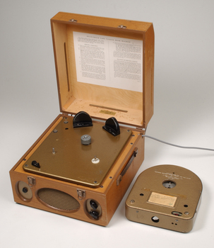 Wooden box with metal grill in front and two clasps.  A golden cartridge sits beside the player.