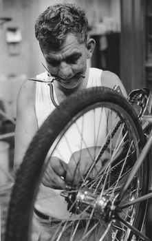 Man working on a Mitchell cycle, holding a pair of pliers in his mouth