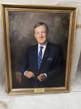 Brown haired man in blue suit, white shirt, blue diagonal striped tie holds a pair of glasses as his elbow rests upon a ledge