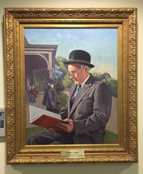Grey haired man in 3 piece grey suit and black bowler sits outside a building looking at a red account book