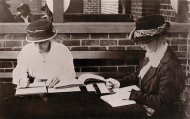 Two women in hats and early 20th century dresses sit as they use a hand frame to guide their stylus as they transcribe 