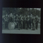 15 members of brass band in matching uniforms stand beside a picket fence
