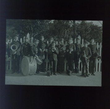 15 members of brass band in matching uniforms stand beside a picket fence