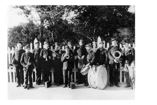 11 members of brass band in matching uniforms with instruments and 1 man beside picket fence