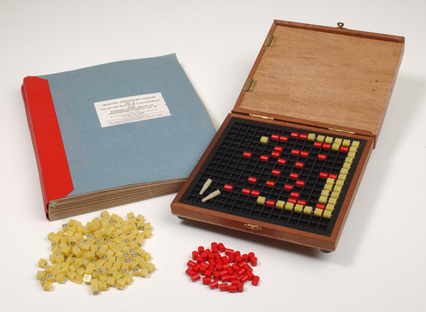 Braille book and wooden box containing yellow and red rubber markers that fit into the grid inset in the box