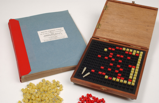 Braille book and wooden box containing yellow and red rubber markers that fit into the grid inset in the box
