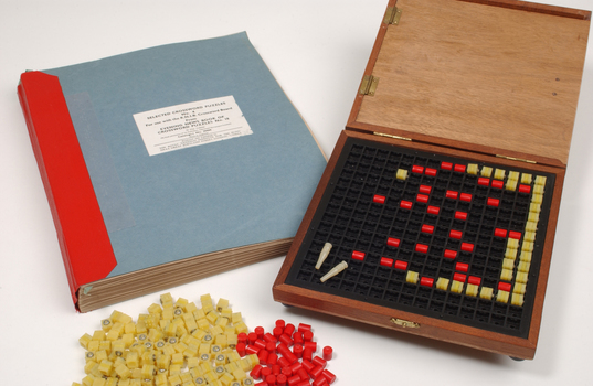 Braille book and wooden box containing yellow and red rubber markers that fit into the grid inset in the box