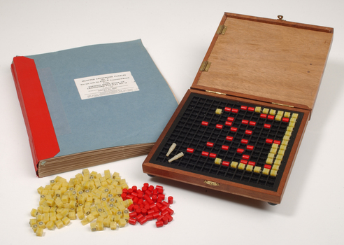 Braille book and wooden box containing yellow and red rubber markers that fit into the grid inset in the box
