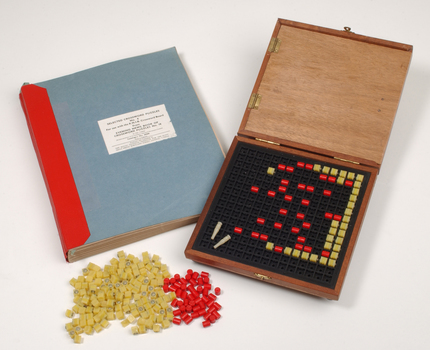 Braille book and wooden box containing yellow and red rubber markers that fit into the grid inset in the box