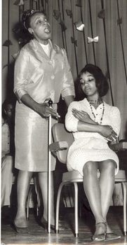 One seated woman pretends to hold a baby whilst another stands behind and sings