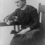Seated woman in dark coloured dress with beading, white gloves and holding a fan and fresh cut roses