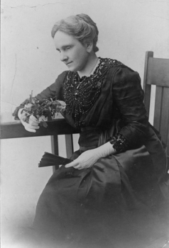 Seated woman in dark coloured dress with beading, white gloves and holding a fan and fresh cut roses