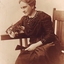 Seated woman in dark coloured dress with beading, white gloves and holding a fan and fresh cut roses in sepia