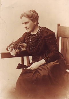 Seated woman in dark coloured dress with beading, white gloves and holding a fan and fresh cut roses in sepia